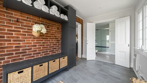 Decoração moderna com parede de tijolos, um banco de madeira escura com cestos, e um corredor que leva a um banheiro.