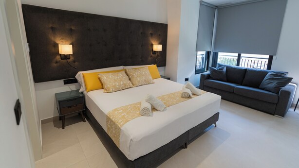 Quarto de hotel moderno com uma cama grande, um sofá cinzento, duas luminárias fixadas na parede e uma varanda com vista.