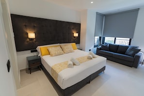 Una habitación de hotel moderna con una cama grande, un sofá gris, dos lámparas fijadas a la pared y un balcón con vistas.