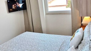 Habitación de hotel con cama, televisión en la pared, ventana con vista a un edificio y equipo de aire acondicionado.
