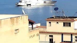 Un yate blanco grande con tres mástiles anclado cerca de un edificio costero con techo de tejas rojas.