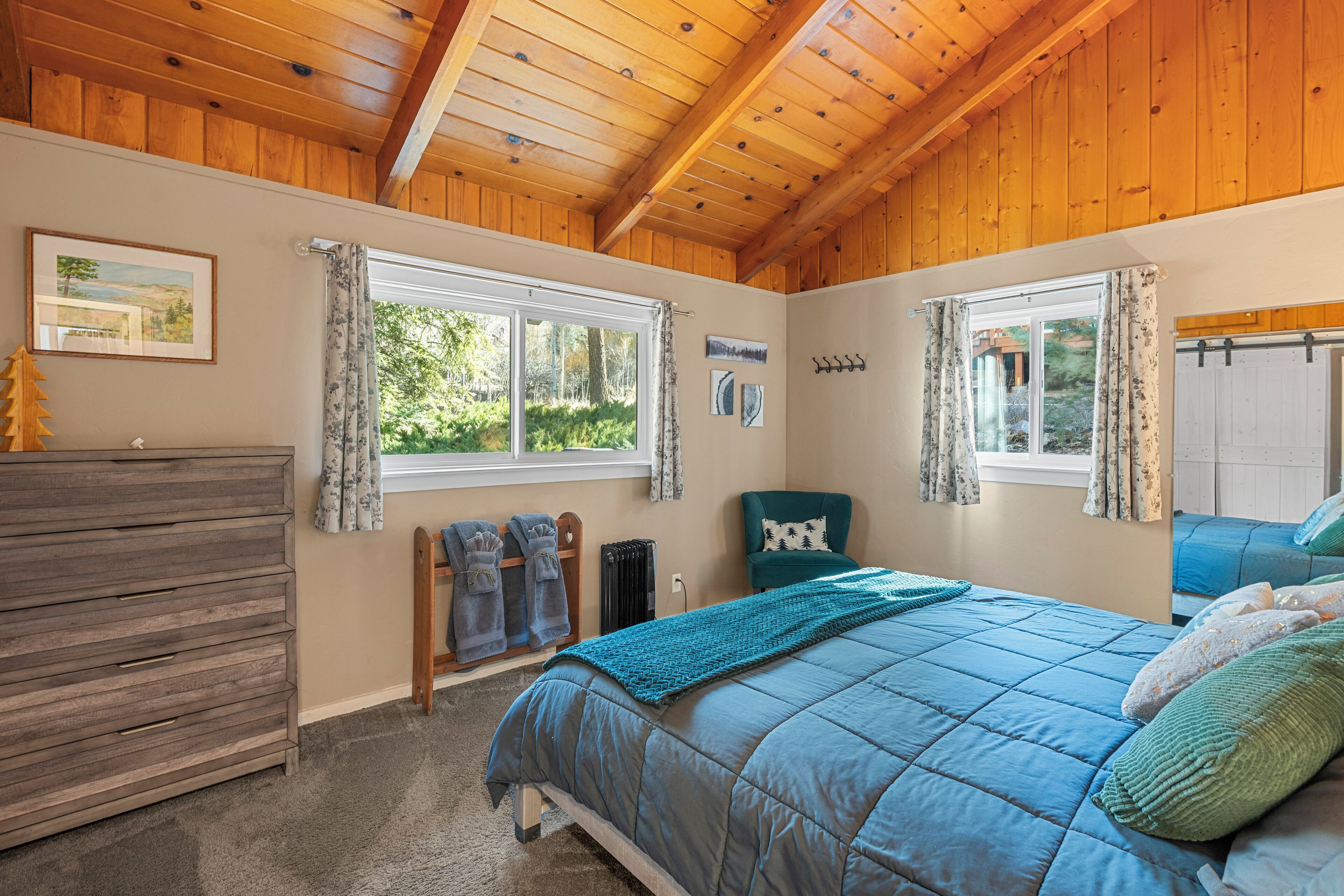 A bedroom with a wooden ceiling, a bed with a blue quilt, a dresser, and a window with curtains.