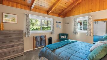 A bedroom with a wooden ceiling, a bed with a blue quilt, a dresser, and a window with curtains.