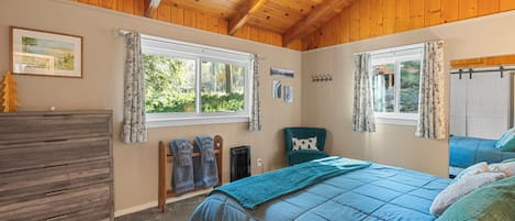 A bedroom with a wooden ceiling, a bed with a blue quilt, a dresser, and a window with curtains.