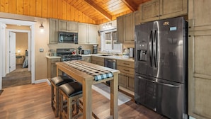 A kitchen with wooden cabinets, a stainless steel refrigerator, and a wooden table with stools. - Peaceful Pines Panorama (Big Bear Lake)