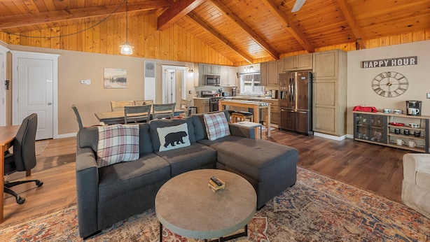 A spacious living room with a sectional sofa, a coffee table, and a dining area with a wooden table and chairs. A kitchen with modern appliances is visible in the background. - Peaceful Pines Panorama (Big Bear Lake)
