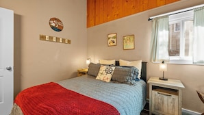 A bedroom with a bed, a nightstand, a window with curtains, a door, and wall decorations. - Peaceful Pines Panorama (Big Bear Lake)