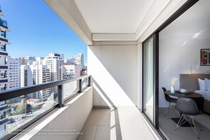 Un balcón moderno con vistas a la ciudad, una zona de comedor con mesa y sillas, y una cama con mesita de noche.