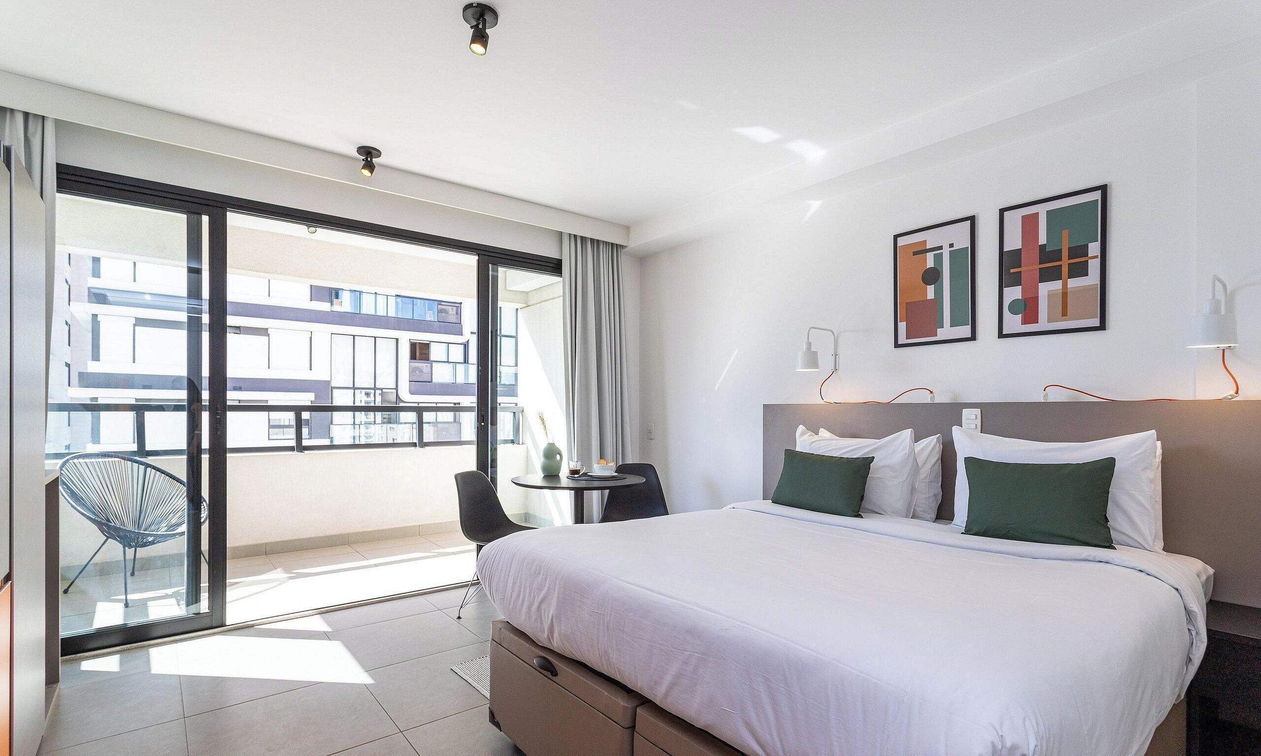 Ein Hotelzimmer mit Bett, Schreibtisch, Stuhl und einem Balkon mit Blick auf Gebäude. Kunstwerke sind an der Wand angebracht.