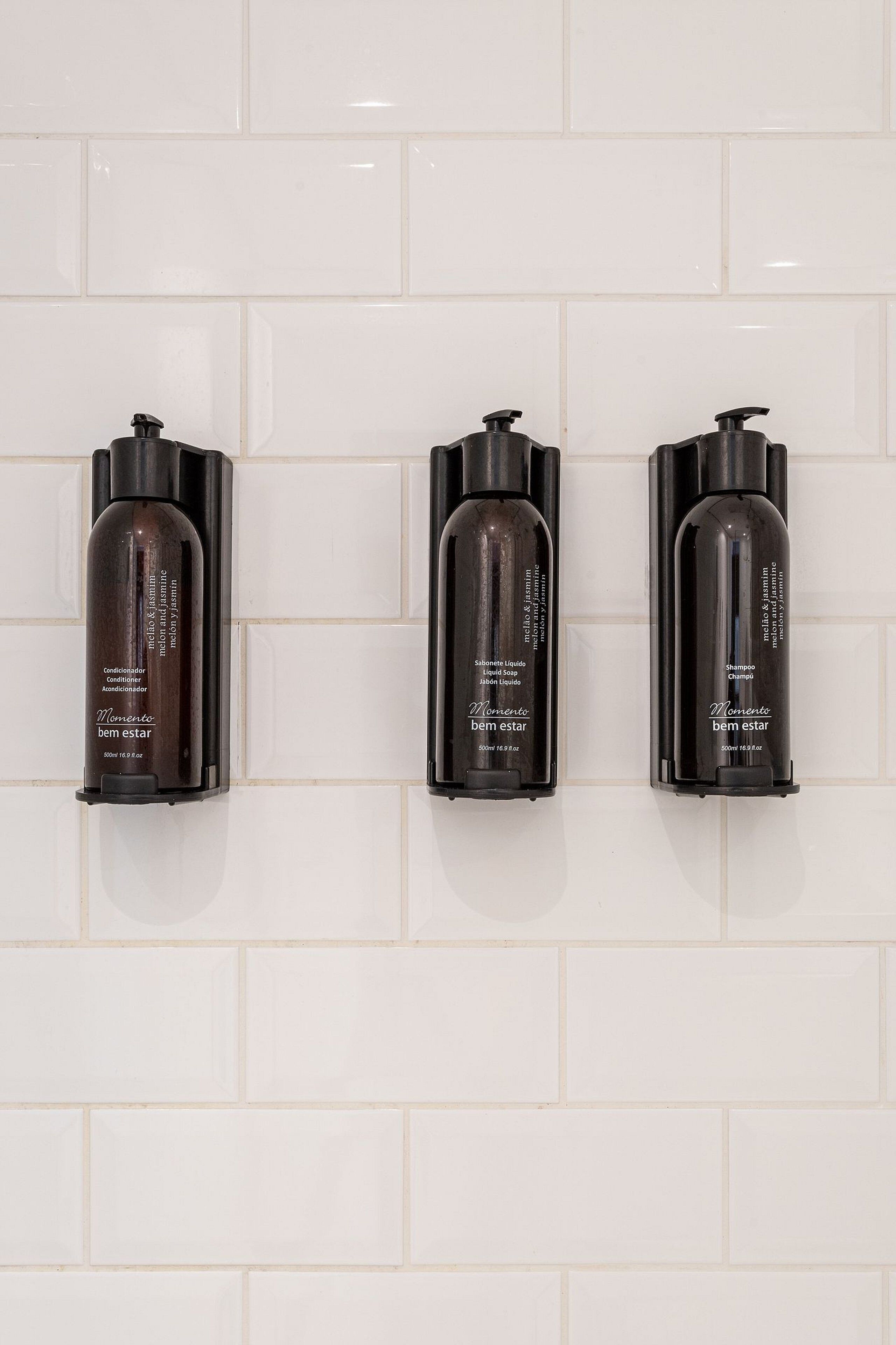 Three bottles of shampoo and conditioner mounted on a white tiled wall.