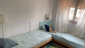 Ein Zimmer mit zwei Einzelbetten, einer Lampe, einem Fenster mit Vorhängen und einem kleinen Tisch.