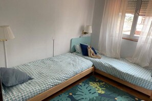 Ein Zimmer mit zwei Einzelbetten, einer Lampe, einem Fenster mit Vorhängen und einem kleinen Tisch.