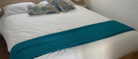 Una cama bien hecha con una manta color aguamarina, almohadas blancas y un cojín estampado con motivos tropicales.