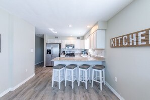 A modern kitchen with white cabinets, a stainless steel refrigerator, a built-in oven, and a bar stool area. - The Sly Fox–Family-Friendly Gulf Escape-Sleeps 20 (Gulf Shores)