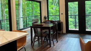 A modern dining area with a wooden table and chairs, large windows with a forest view, and a mounted television. - NEW The Outlook (Laurelville)