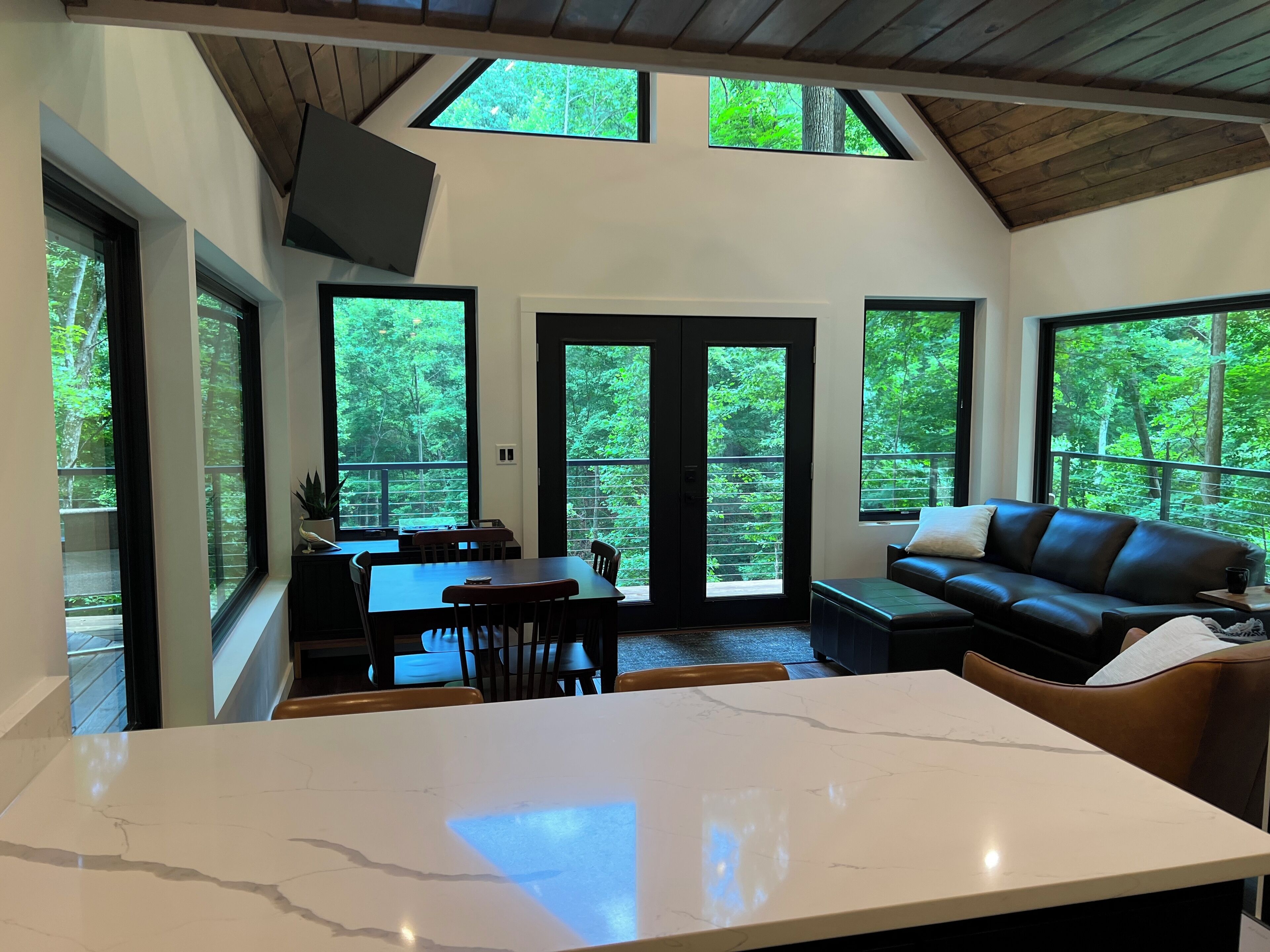 A modern interior with a large white marble kitchen island, dining area with wooden chairs, a black leather sofa, and large windows offering a view of a forested area.