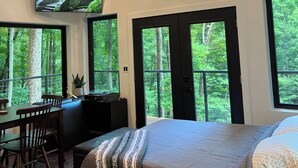 A bedroom with a large bed, a desk, and a chair. There are large windows with a view of trees. - NEW The Outlook (Laurelville)