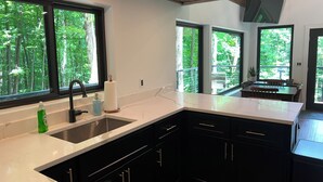 A modern kitchen with a large island, dark cabinetry, and large windows offering a view of the forest. - NEW The Outlook (Laurelville)
