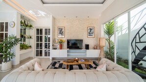 A modern living room with a large sofa, a flat-screen TV, and a coffee table. There are potted plants and a staircase visible in the background. - Peaceful Retreat-2BR,Canggu (Mengwi)