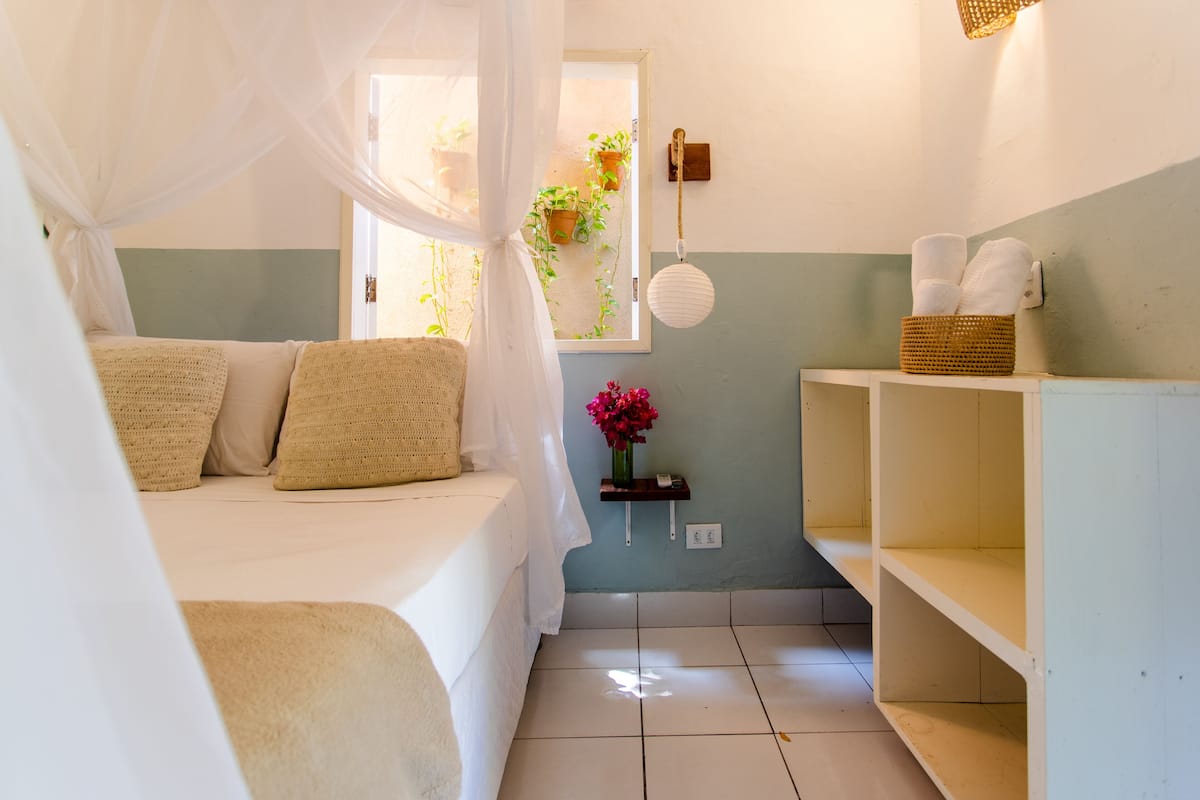 A bedroom with a bed, a bedside table, a window with plants, and a wall-mounted shelf with towels.