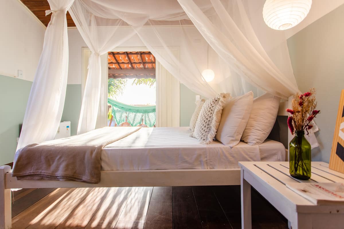 A bedroom with a bed, white curtains, a bedside table with a vase, and a view of a green hammock outside.