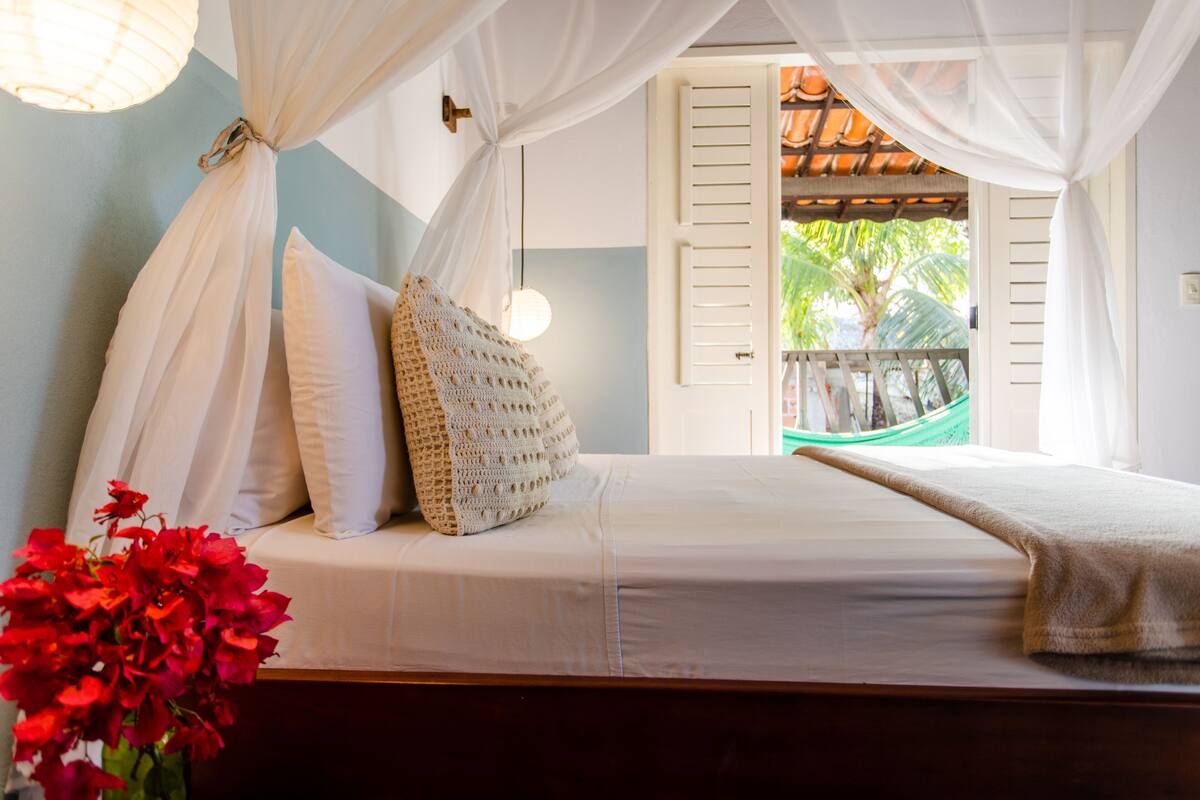 A bed with white bedding, a crocheted pillow, and a view of a balcony with a hammock.