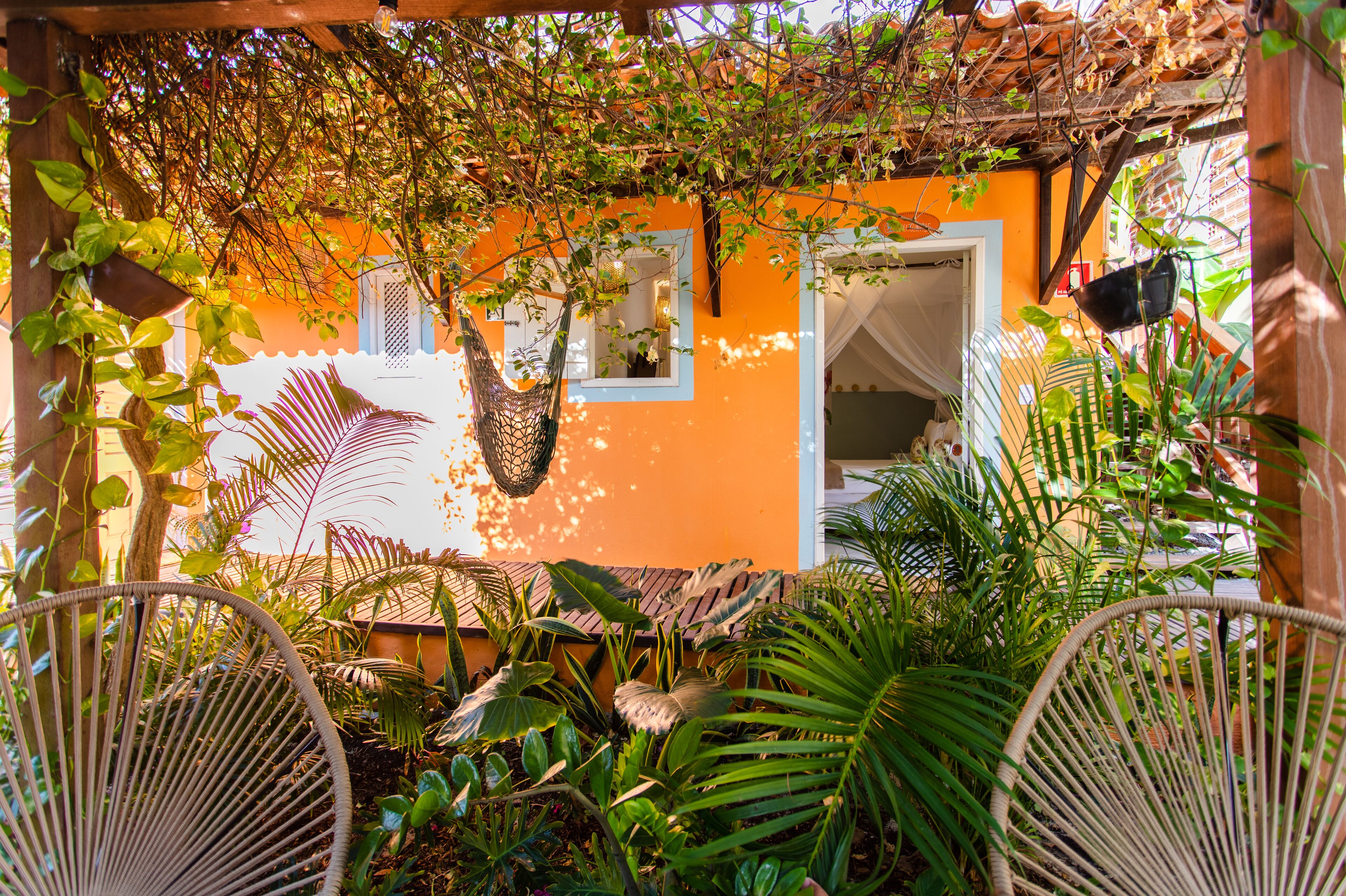 Cómodo espacio al aire libre con sillas de mimbre, rodeado de exuberante vegetación tropical y un edificio naranja vibrante con ventana y puerta.