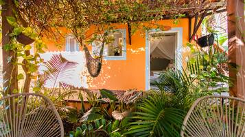 Cómodo espacio al aire libre con sillas de mimbre, rodeado de exuberante vegetación tropical y un edificio naranja vibrante con ventana y puerta.