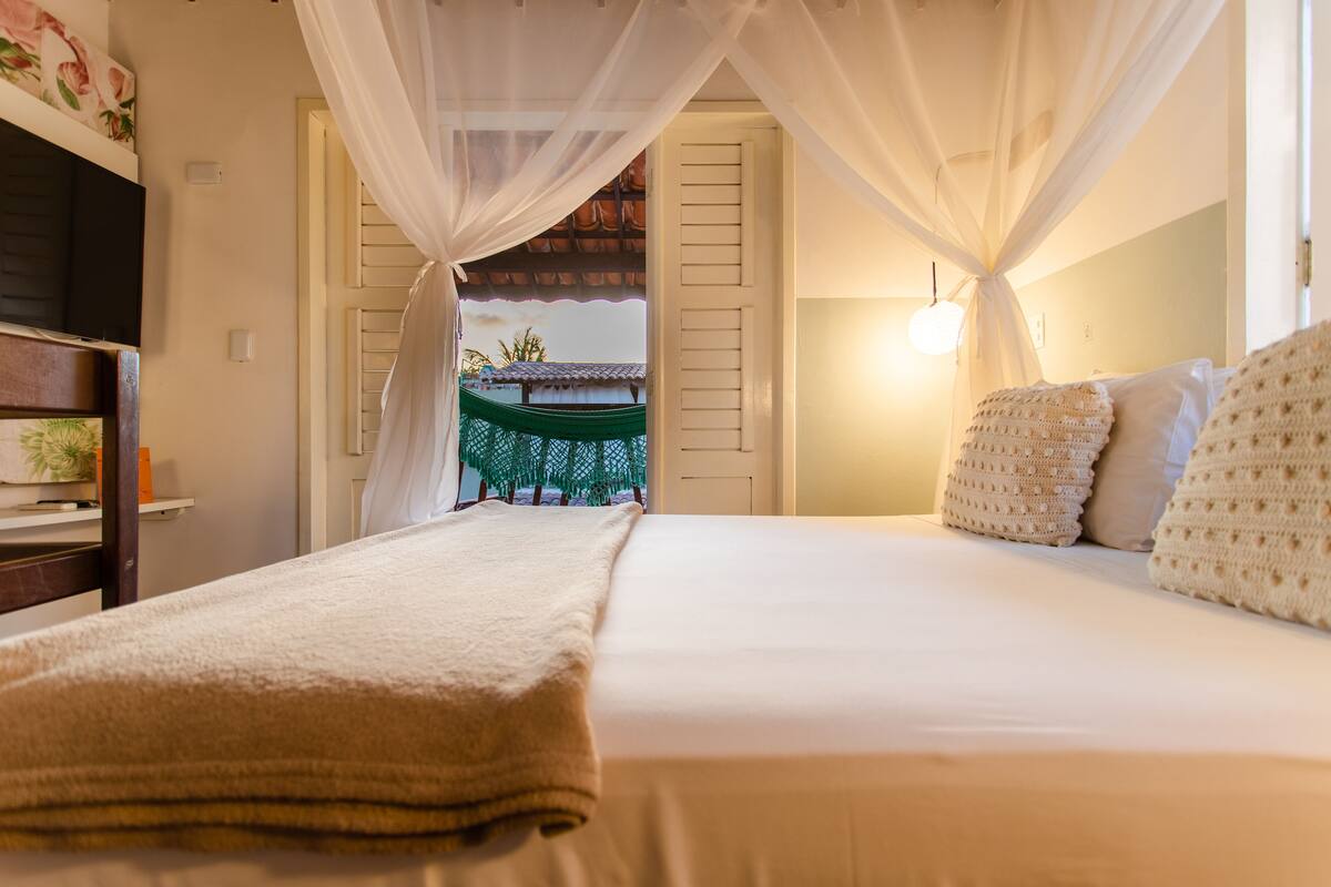A bedroom with a bed, white curtains, a bedside lamp, and a view of a traditional building outside.