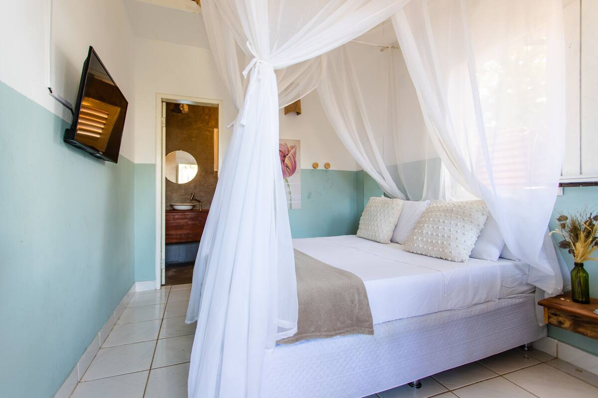 A bedroom with a bed under a white canopy, a bedside table with a vase, and a wall-mounted light.
