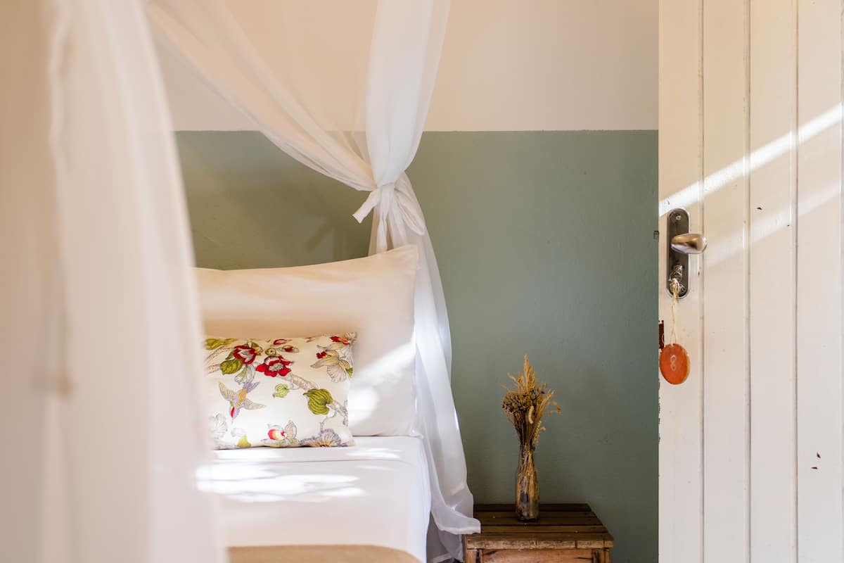 A bed with a canopy, a floral pillow, a vase with dried flowers, and a wooden nightstand.