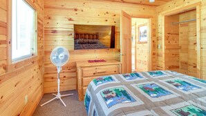 Un dormitori de cabana de fusta amb un llit, una petita estant, un ventilador i una finestra.