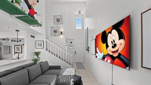 A modern living room with a grey sofa, a flat-screen TV displaying a cartoon character, and a staircase leading to another level. - My Disney Home (Kissimmee)
