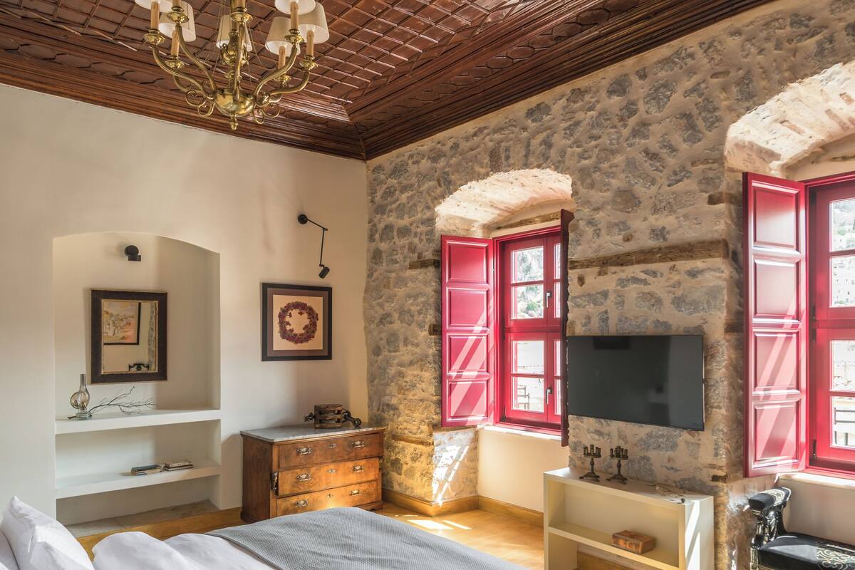 Un dormitorio con techo de madera, paredes de piedra, una cama, un aparador, un televisor y contraventanas rojas.