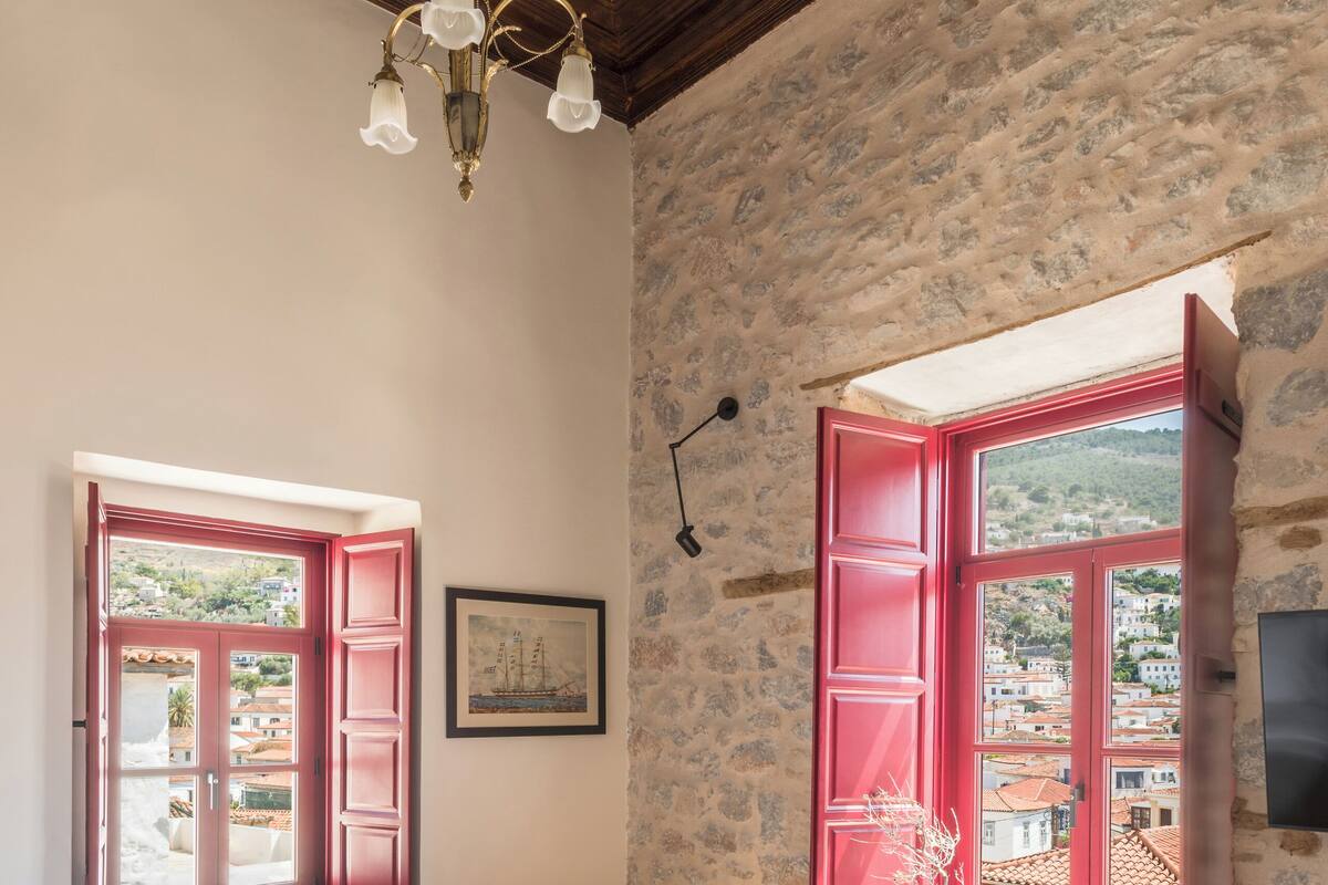 Un dormitorio con techo de madera, cama con ropa de cama blanca, dos ventanas con contraventanas rojas, pared de piedra y una pequeña mesa con un libro.