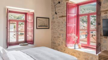 Un dormitorio con techo de madera, cama con ropa de cama blanca, dos ventanas con contraventanas rojas, pared de piedra y una pequeña mesa con un libro.
