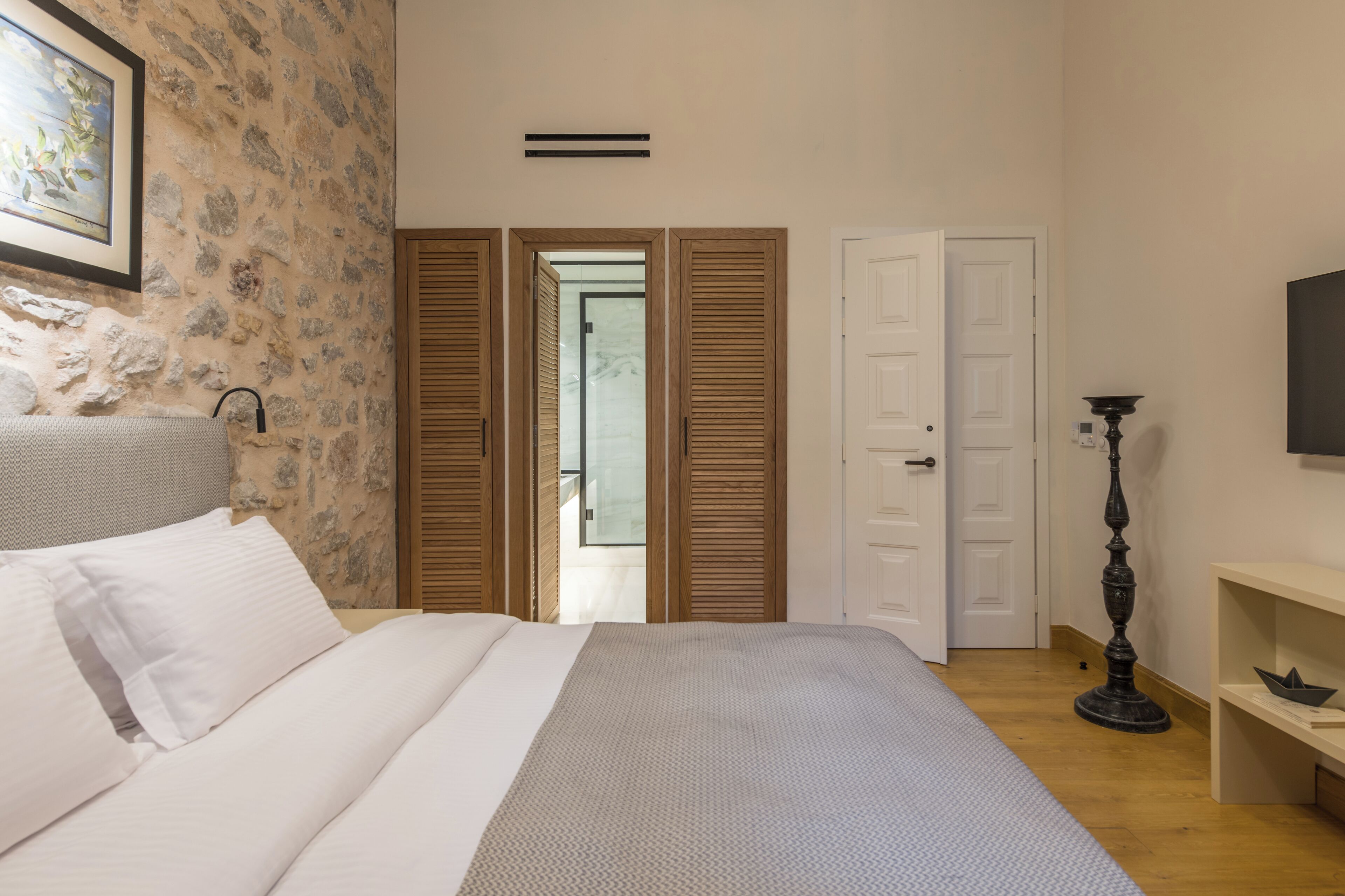 Quarto com cama, guarda-roupa de madeira, porta, luminária de chão e televisão fixada na parede.