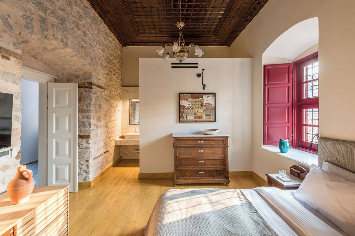 Un dormitorio con pared de piedra, techo de madera, cama, cómoda, ventana con contraventanas rojas y un cuadro en la pared.