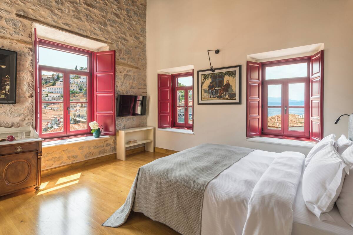 Un dormitorio con una cama grande, un aparador de madera, un ventanal con contraventanas rojas, una mesita con un florero y un cuadro en la pared.