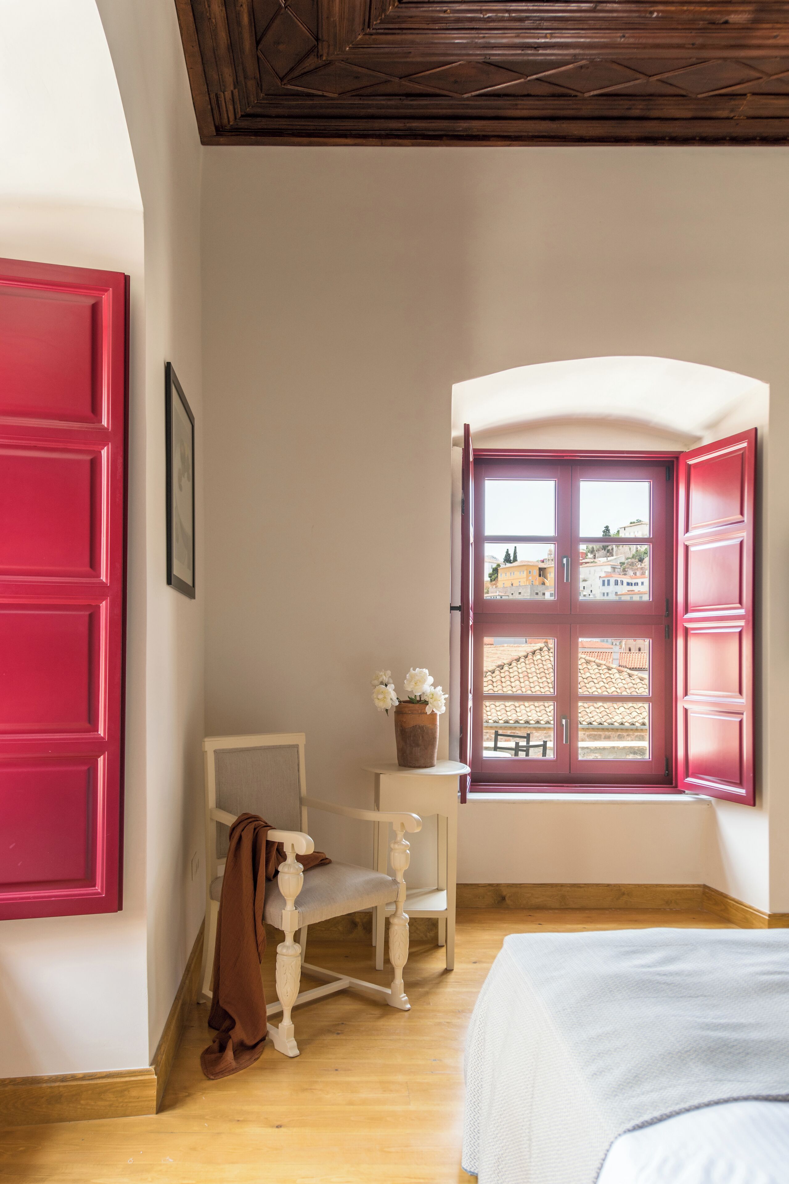 Ein Zimmer mit Holzboden, einem weißen Bett, einer roten Tür und einem Fenster mit roten Lamellen.