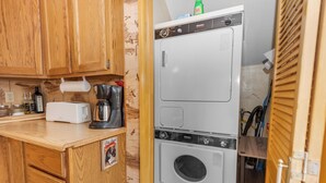 A compact kitchen with wooden cabinets, a white toaster, a coffee maker, and a stacked washer and dryer. - Boulder Pines (Big Bear Lake)