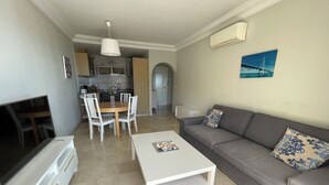A modern living room with a grey sofa, a white coffee table, and a TV on a white cabinet. There is a kitchen area with white cabinets and a dining table with chairs. - Lovely! Close to marina, beaches and town (Estepona)