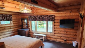 Ein Schlafzimmer in einer Blockhütte mit Bett, Schminktisch, Kommode, Bank und einem an der Wand befestigten Fernseher.