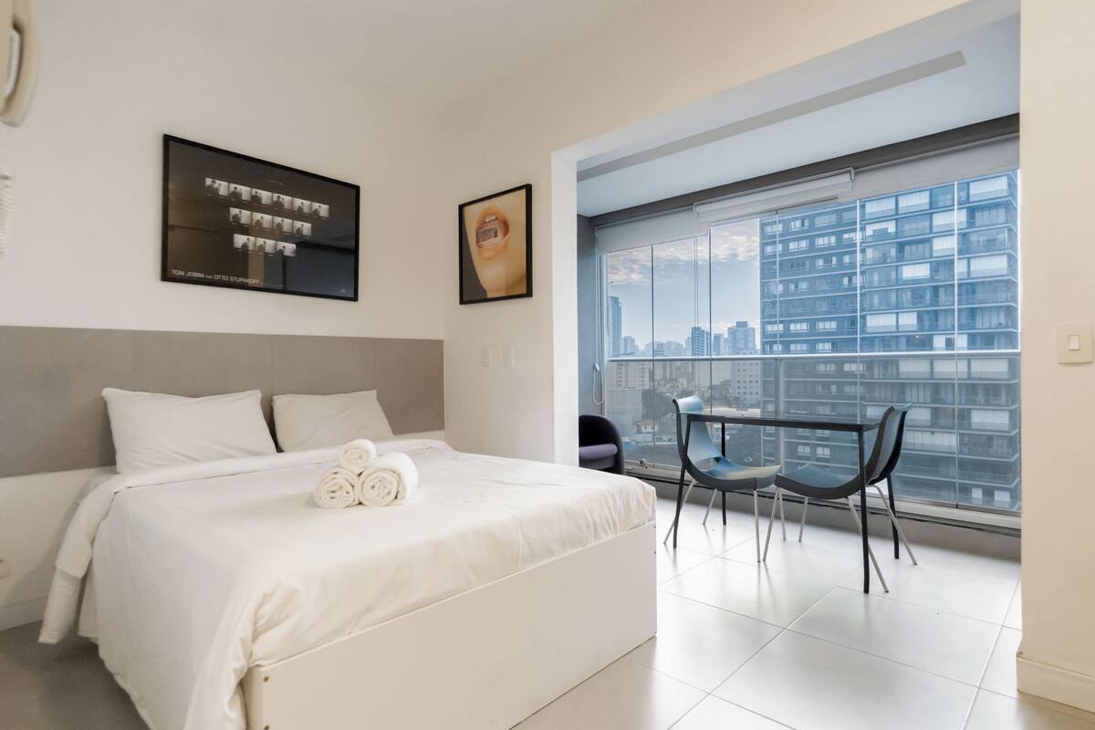 Quarto de hotel moderno com uma cama grande, vista para a cidade através de uma janela do chão ao teto, e uma pequena varanda com uma cadeira.