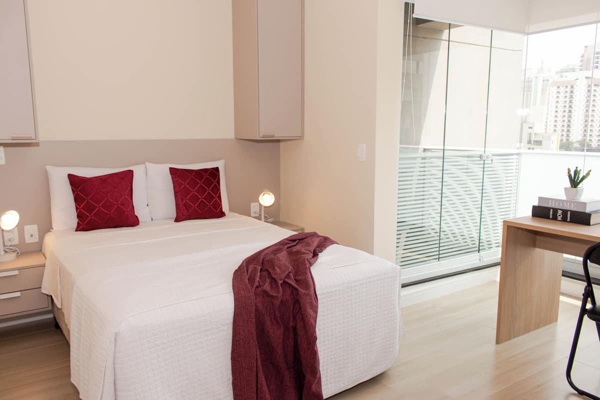 Quarto moderno com uma cama grande, uma escrivaninha e vista da cidade através de amplas janelas.