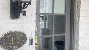 Un b âtiment en briques blanches avec une porte gris foncé, une lanterne noire au-dessus de la porte et une plaque portant l’inscription : « Hilltop Cottage, Mockingbird Lane 3105 ».