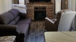 A living room with a stone fireplace, a flat-screen TV mounted above it, a dark-colored sofa, a light-colored armchair, and a patterned throw blanket. - Cozy tiny home w/lofts deck grill hammock (Phenix City)