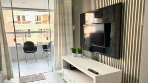 Un salón moderno con un televisor de pantalla plana montado en una pared blanca, un mueble blanco para equipos con un control remoto, y un balcón con sillas y vistas a edificios.
