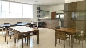 Un comedor amplio con varias mesas y sillas, una cocina equipada con electrodomésticos modernos, y ventanas grandes que permiten la entrada de luz natural.
