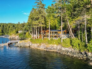 A lakeside cabin with a dock, surrounded by dense forest and a clear sky. - Kingfisher Manor (Sooke)